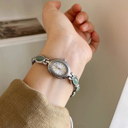Hand wearing a silver watch with green gemstones on a neutral background