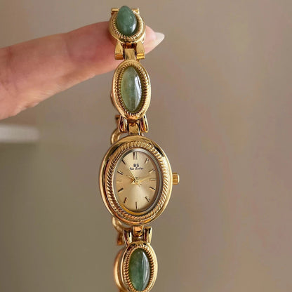 Gold watch with green gemstones held by a hand against a neutral background