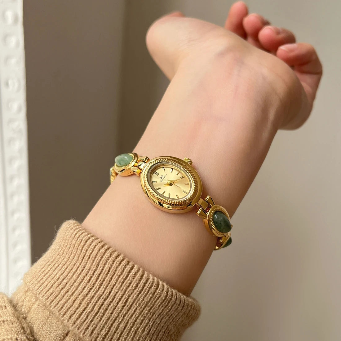 Gold wristwatch with green accents on a wrist, wearing a beige sleeve.