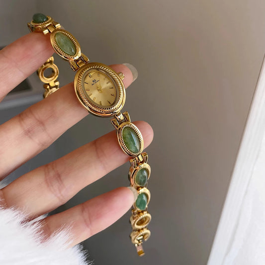Gold watch with green gemstones held by a hand against a neutral background